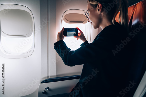 Millennial passenger using smartphone camera for shooting video vlog from window porthole in jetliner airplane during international comfortable flight in business class, woman photographing view