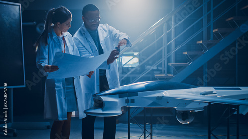 Two Aerospace Engineers Work On Unmanned Aerial Vehicle / Drone Prototype. Aviation Scientists in White Coats Holding Blueprints. Laboratory with Commercial Aerial Surveillance Aircraft photo
