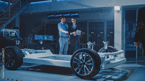 Auto Industry Design Facility: Male Chief Engineer Shows Car Prototype to Female Car Designer. Electric Vehicle Platform Chassis Concept with Wheels, Engine and Battery. © Gorodenkoff