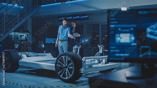 Auto Industry Design Facility: Male Chief Engineer Shows Car Prototype to Female Car Designer. Electric Vehicle Platform Chassis Concept with Wheels, Engine and Battery. © Gorodenkoff