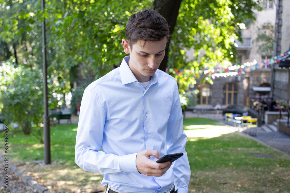 Portrait of young man in a blue shirt on the city with a phone in his hand looks at phone with a trees on the background. The man in conversation. American or European appearance. Summer day. Portrait of young man in a blue shirt on the city with a phone in his hand looks at phone with a trees on the background. The man in conversation. American or European appearance. Summer day.