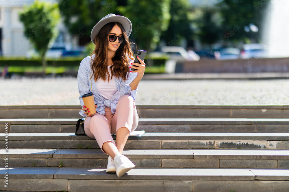 Beautiful young girl looking at her phone and smiling sitting on stairs in urban background.