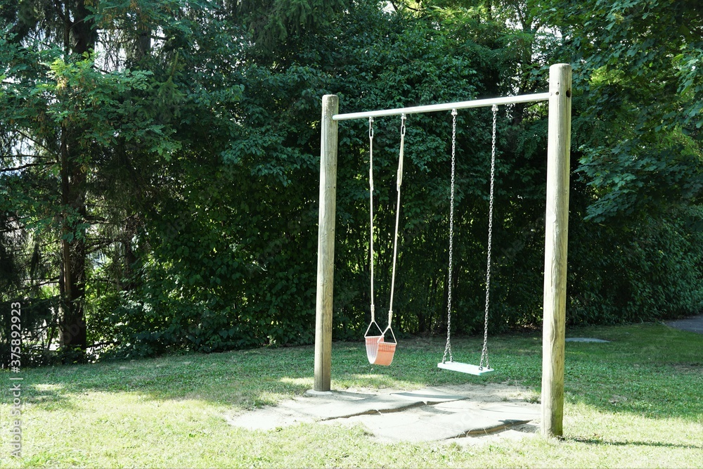 Double swing frame with two sets of swing for children from cloth and plastic. Frame is constructed from round timbers and metal bar. Swing is in garden surrounded by trees and bushes.