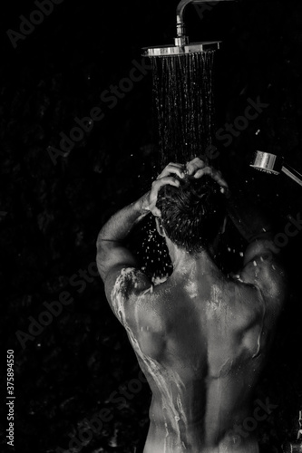 Topless handsome man taking a shower washing hair with shampoo product under water falling from luxury outdoor rain shower head.