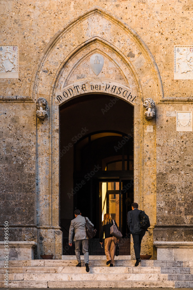 Three bankers are entering the headquarter of the Banca Monte dei Paschi di Siena, one of the oldest banks in the world, through the main door. The building is the famous Palazzo Salimbeni.