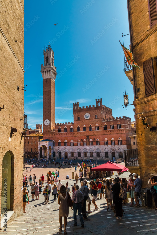 Picturesque view of the town hall Palazzo Pubblico with the complete bell tower Torre del Mangiaa and the Cappella di Piazza; taken on the sloped Costa Barbieri street between two buildings in Siena.
