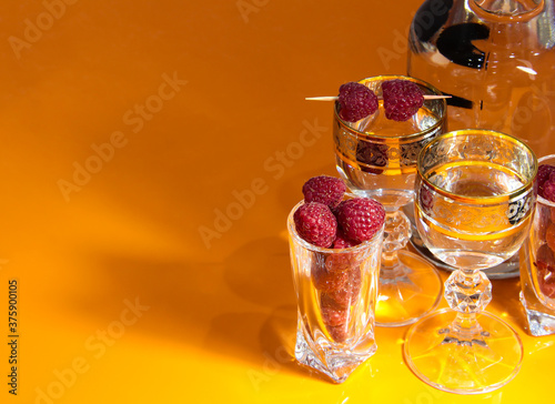 A bottle and glasses of alcohol and raspberries on the orange table.