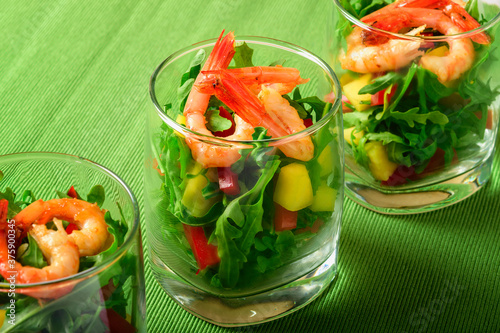 A salad of mango, sweet pepper, rukkola and garlic-fried shrimp in a glass. Close up. Selective focus. Green background.