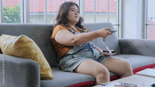 The overweight young woman sitting on the sofa in the living room, turn off TV and eat chocolate cake, she asleep at the end. Chill and have fun at home. Day off at home concept.