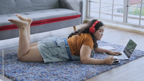 Young woman overweight Lie down on the carpet in the living room, using laptop and wearing red headphone and singing and have the chocolate cake nearby. Look chill and fun at home.