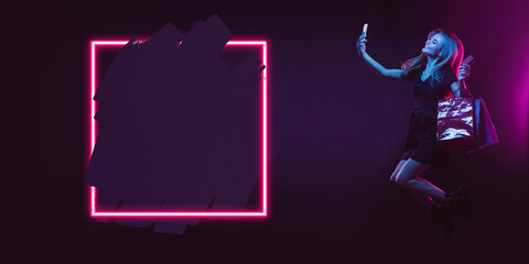 Selfie, jumping. Portrait of young woman in neon on dark studio backgound. Human emotions, black friday, cyber monday, purchases, sales, finance concept. Copyspace. Seamless post for instagram.