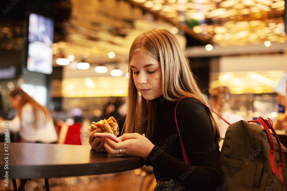 A beautiful teenage girl sitting in a cafe looks at the phone and eats a hamburger. Happy child eating junk food in a restaurant.