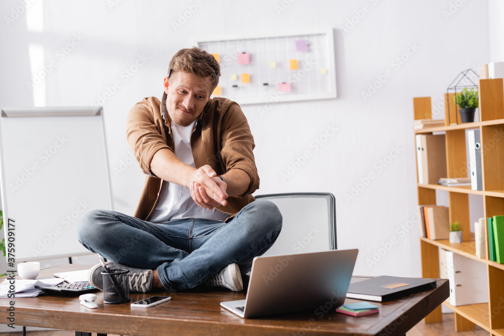 Selective focus of young businessman gesturing while sitting near laptop and stationery on office table Selective focus of young businessman gesturing while sitting near laptop and stationery on office table