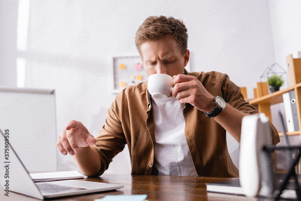 Selective focus of businessman drinking coffee and pointing at laptop in office Selective focus of businessman drinking coffee and pointing at laptop in office