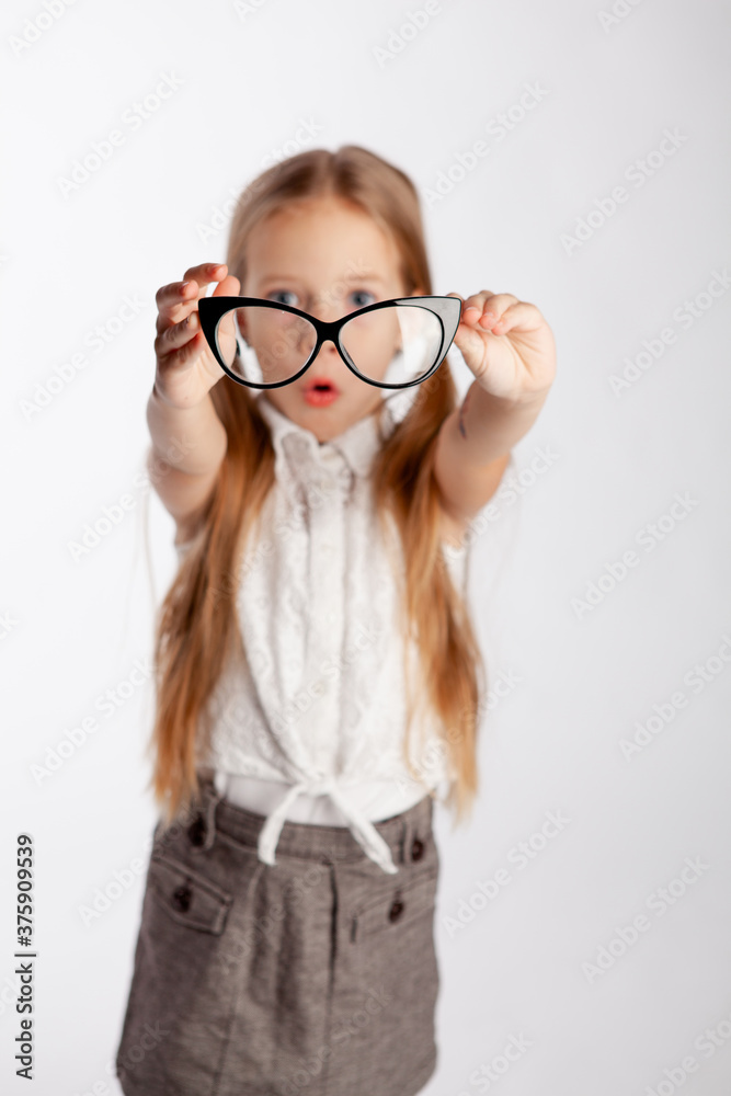 little girl in a strict gray skirt and white blouse holds glasses in outstretched arms. Glasses in focus, the girl is out of focus