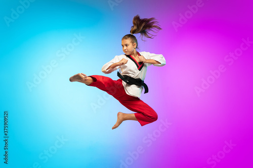 In jump. Karate, taekwondo girl with black belt isolated on gradient background in neon light. Little caucasian model, sport kid training in motion and action. Sport, movement, childhood concept.