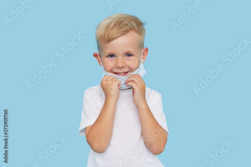 End of quarantine. Portrait of cute little child boy removes the protective medical mask from the face on blue background. Virus protection and healthcare concept.