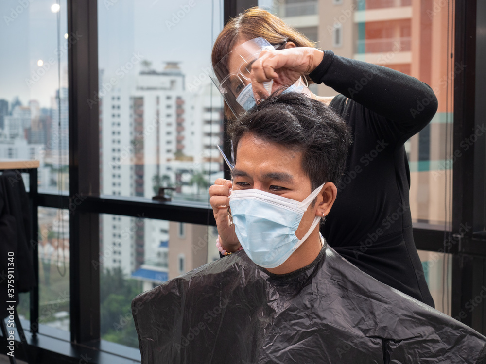 Asia female hairdresser takes hair cut to a caucasian man both are wearing a medical mask to protect themselves against coronavirus. New normal concept..