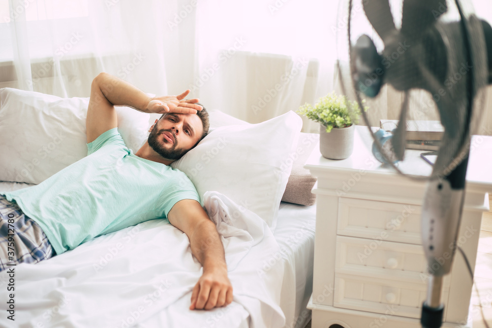 Sweaty young man is trying to refresh from the heat with a fan while lying in bed at home. Sweaty young man is trying to refresh from the heat with a fan while lying in bed at home.