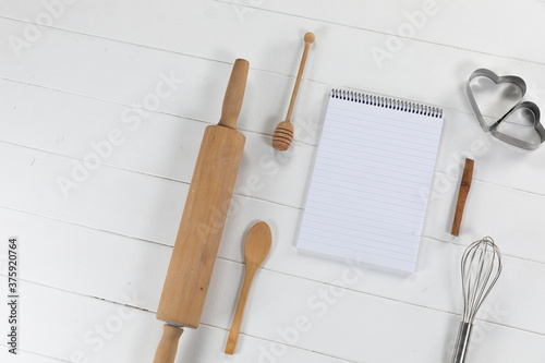 View of a composition of a notebook with cookie cutters, whisker and rolling pin on white surface