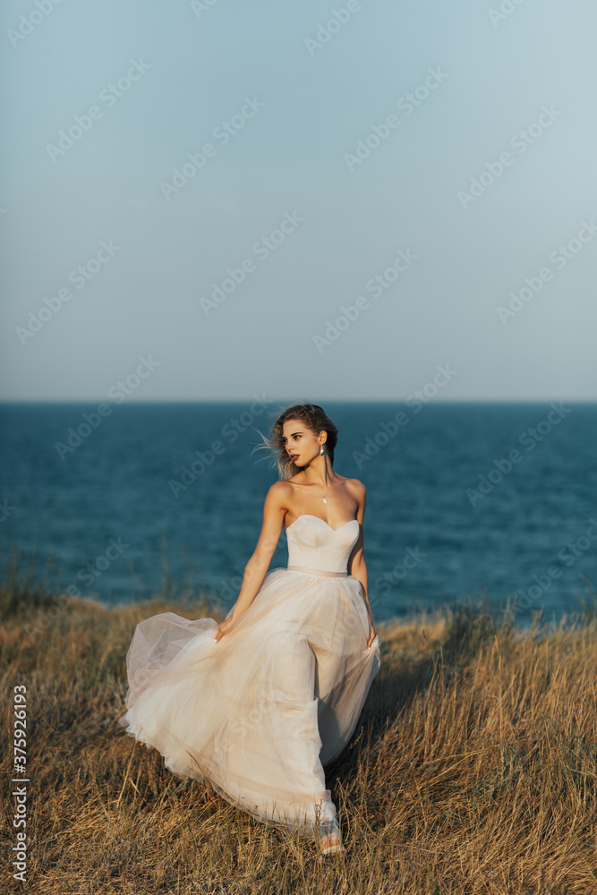 A young beautiful girl in a long pale rose dress walks along the beach against the background of the  blue sea. Copy space.