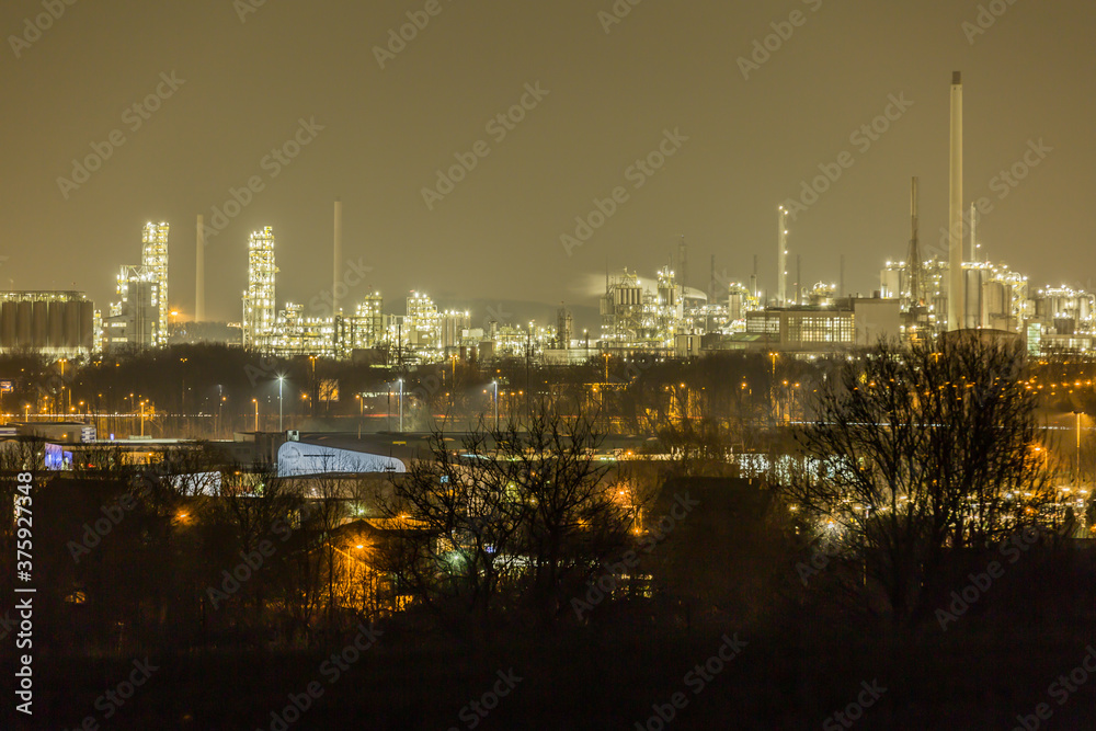 Industrial complex with its metal structures illuminated by white lights seen from a hill with bare trees, quiet night in southern Limburg in the Netherlands Holland