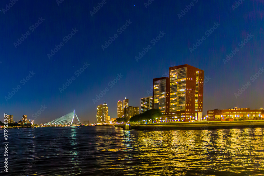 Obraz premium Night cityscape in Rotterdam with its buildings with lights on and the Erasmus cable-stayed bridge over the Maas river, clear blue sky in the Netherlands