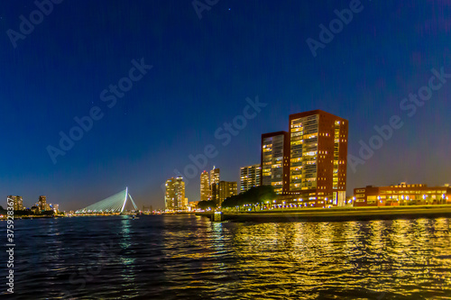Night cityscape in Rotterdam with its buildings with lights on and the Erasmus cable-stayed bridge over the Maas river, clear blue sky in the Netherlands