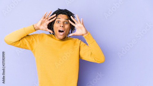 Young black man wearing rasta hairstyle receiving a pleasant surprise, excited and raising hands.