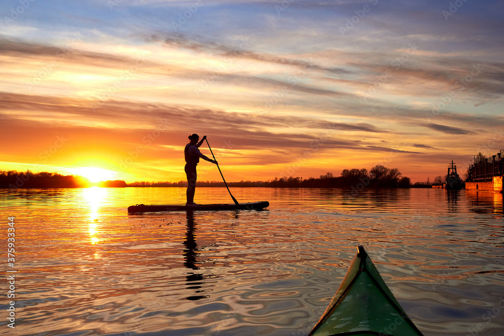 View from bow of kayak on silhouette of woman paddling on a SUP boards on the autumn Danube river at sunset. Stand up paddle boarding. Rear view. Backlight.