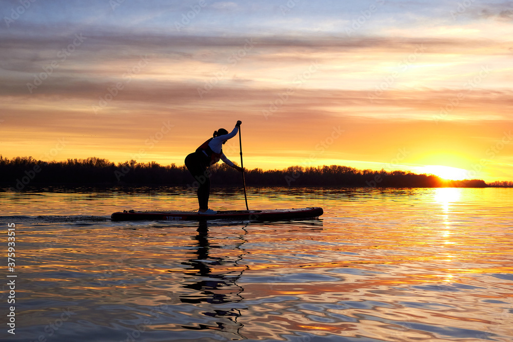 Silhouette of woman paddle on stand up paddle boarding (SUP) on quiet winter or autumn river at sunset. Colorful sunset over the river