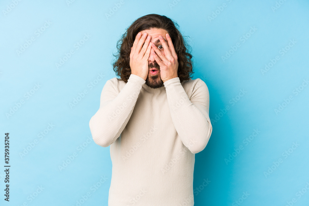 Young long hair man isolated on a blue background blink through fingers frightened and nervous. Young long hair man isolated on a blue background blink through fingers frightened and nervous.