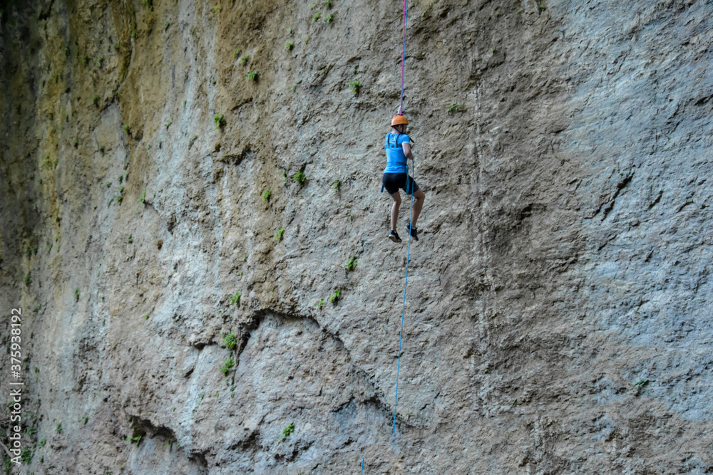 A rock climber in full uniform is going down a gray rock. The athlete climbs the rock with the help of the ropes.