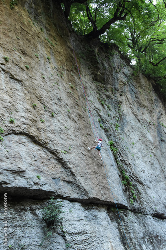 A rock climber in full uniform is going down a gray rock. The athlete climbs the rock with the help of the ropes.