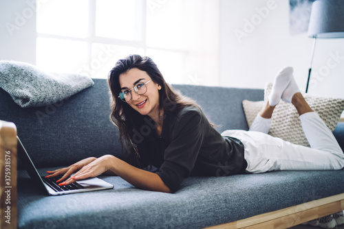 Portrait of smiling caucasian female in eyewear for vision correction typing on laptop computer share content in social networks, cheerful woman looking at camera working remotely on sofa at home