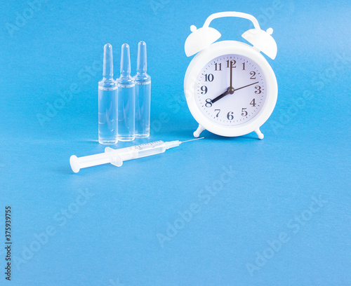 White alarm clock, medical ampoules, syringe on a blue background with copy space. The concept of vaccination against infection and viruses