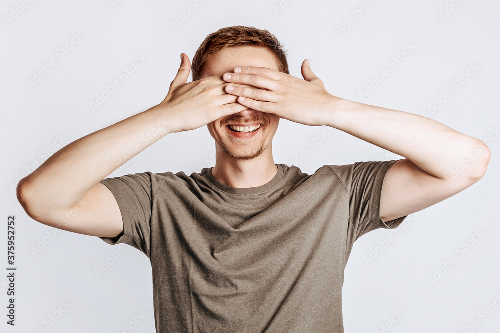 Handsome young man closes his eyes with two hands and smiles. Brunette with a beard in a green shirt posing on a gray background. Place for advertising. I do not see. Handsome young man closes his eyes with two hands and smiles. Brunette with a beard in a green shirt posing on a gray background. Place for advertising. I do not see.