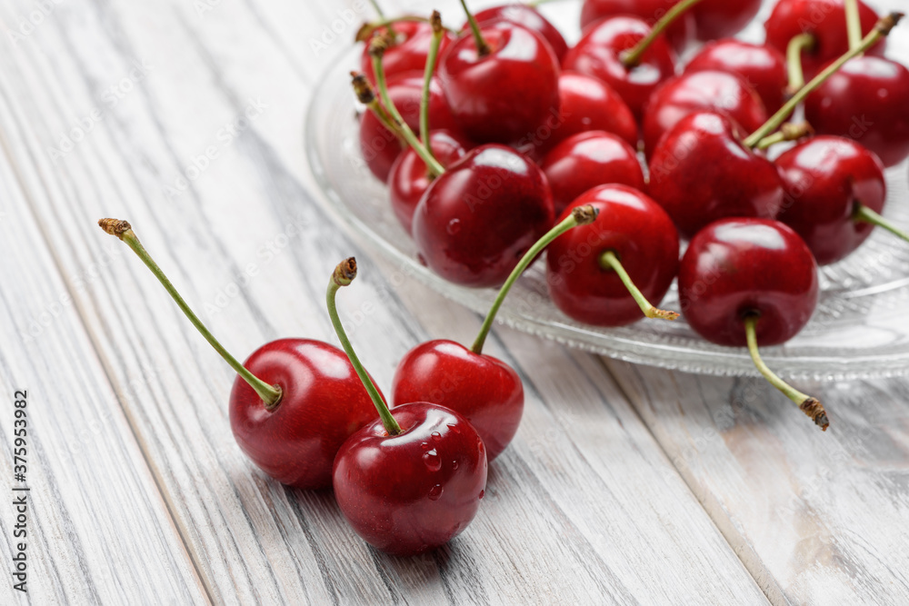 Fresh cherry berries with drops of water in a glass plate on a white wooden table. Close-up macro shot Fresh cherry berries with drops of water in a glass plate on a white wooden table. Close-up macro shot