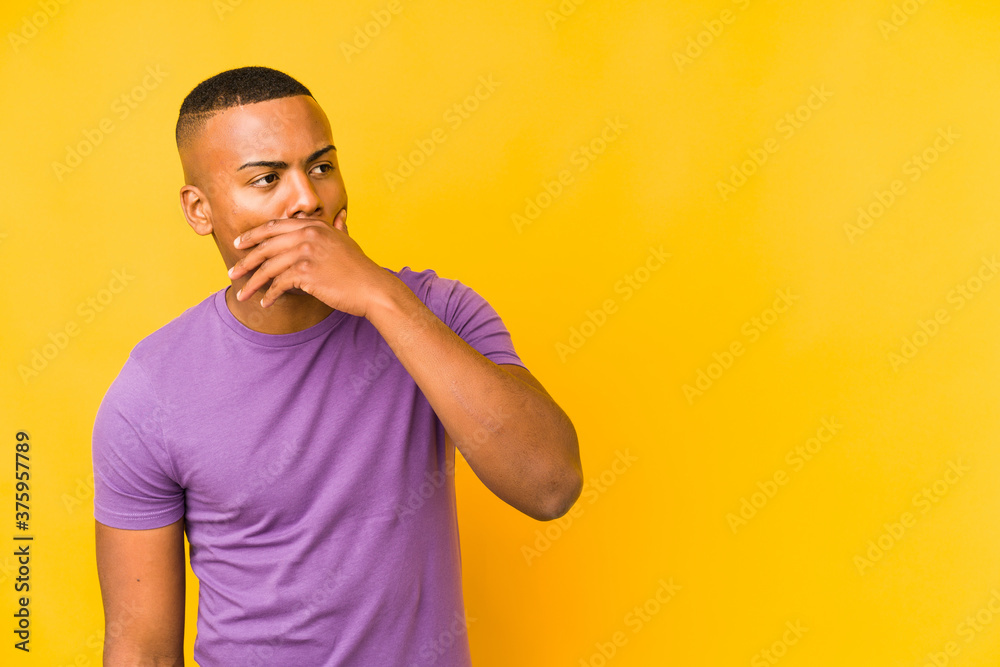 Young latin man isolated on yellow background thoughtful looking to a copy space covering mouth with hand. Young latin man isolated on yellow background thoughtful looking to a copy space covering mouth with hand.