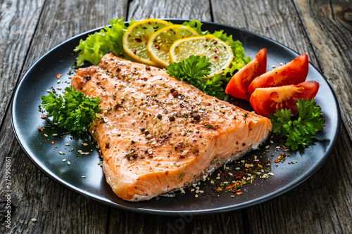 Fried salmon fillet with lettuce, lemon and tomatoes served on black plate on wooden table 