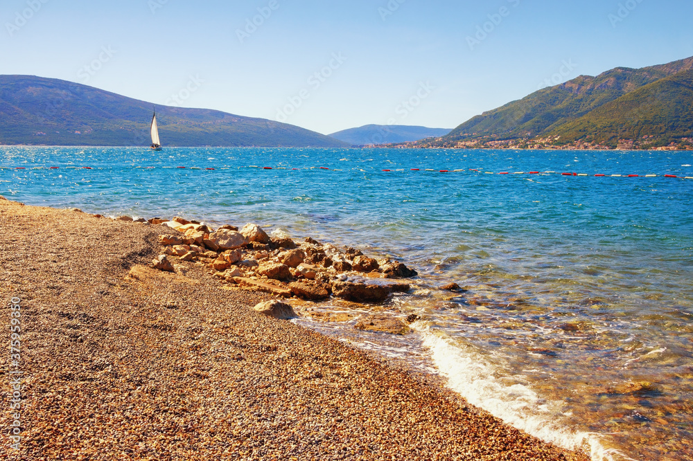 Autumn vacation concept. Calm sunny day, picturesque Mediterranean landscape, deserted beach and white sailboat on water.  Montenegro, Adriatic Sea, view of Bay of Kotor near Tivat city