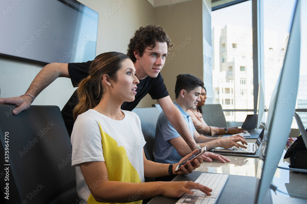 Young woman using computer in office typing on keyboard and checking mobile phone, showing male co-worker something on her screen with co-workers working in background Young woman using computer in office typing on keyboard and checking mobile phone, showing male co-worker something on her screen with co-workers working in background