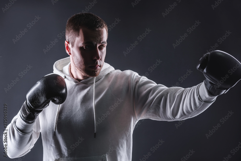 Young man in boxing gloves and a white sweatshirt on a dark background in the studio. The concept of victories and battles