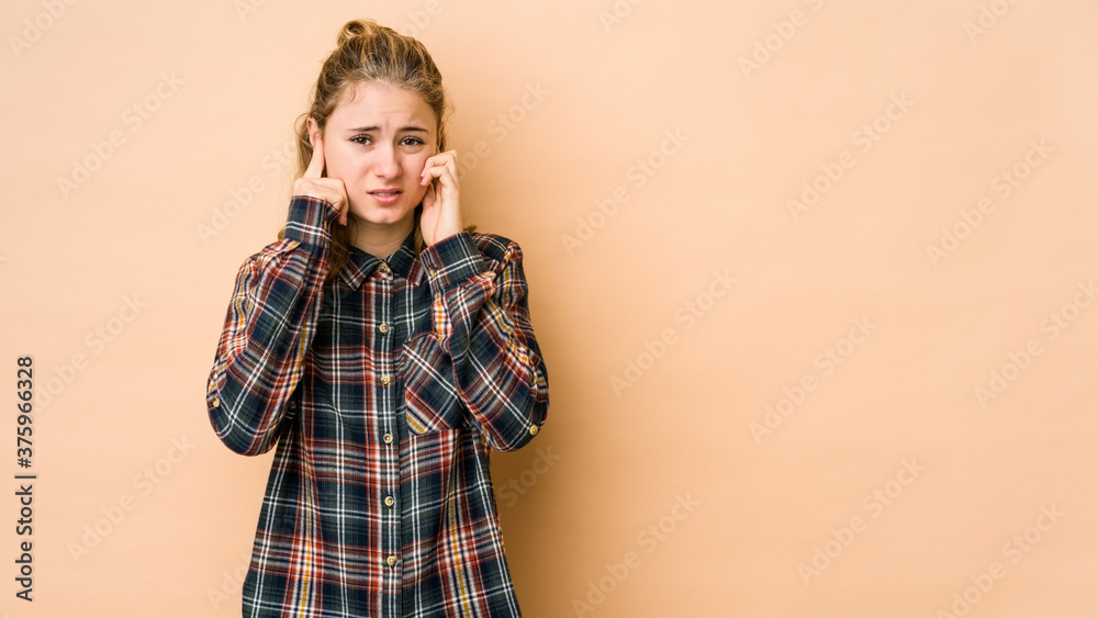 Young caucasian woman isolated on beige background covering ears with fingers, stressed and desperate by a loudly ambient. Young caucasian woman isolated on beige background covering ears with fingers, stressed and desperate by a loudly ambient.