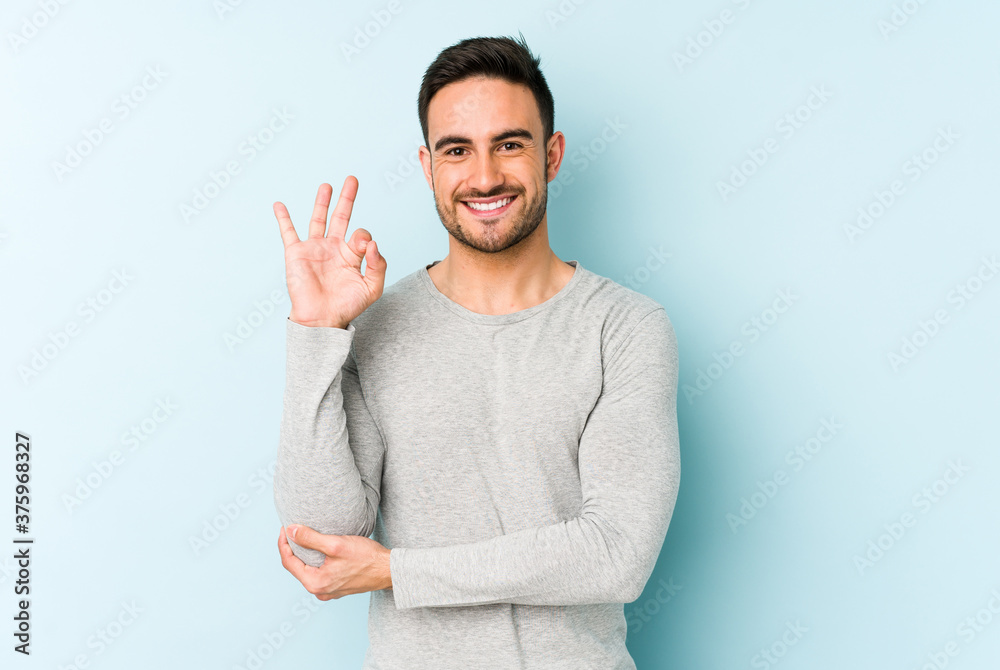 Young caucasian man isolated on blue background winks an eye and holds an okay gesture with hand. Young caucasian man isolated on blue background winks an eye and holds an okay gesture with hand.