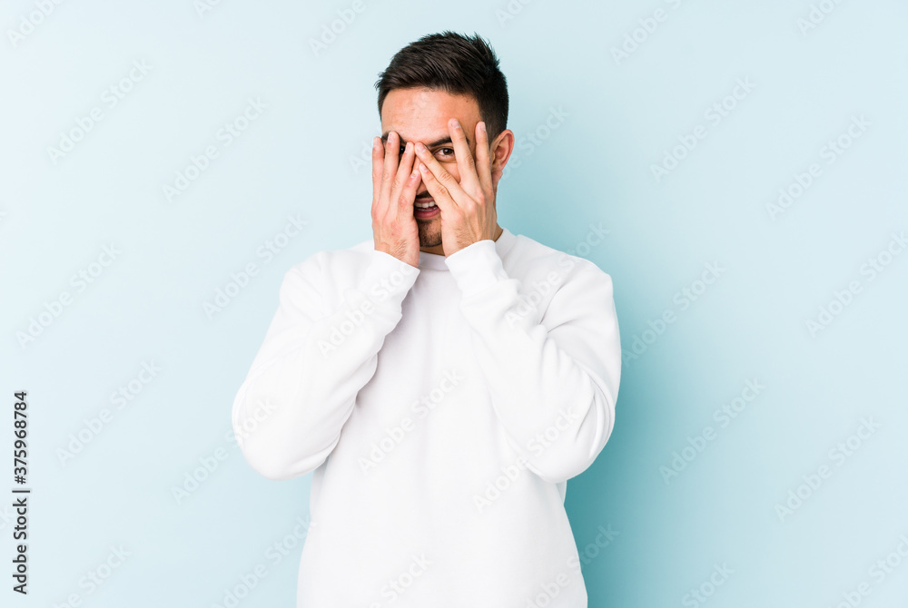 Young caucasian man isolated on blue background blink at the camera through fingers, embarrassed covering face. Young caucasian man isolated on blue background blink at the camera through fingers, embarrassed covering face.