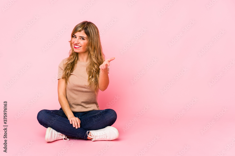 Young blonde caucasian woman sitting on a pink studio stretching hand at camera in greeting gesture. Young blonde caucasian woman sitting on a pink studio stretching hand at camera in greeting gesture.