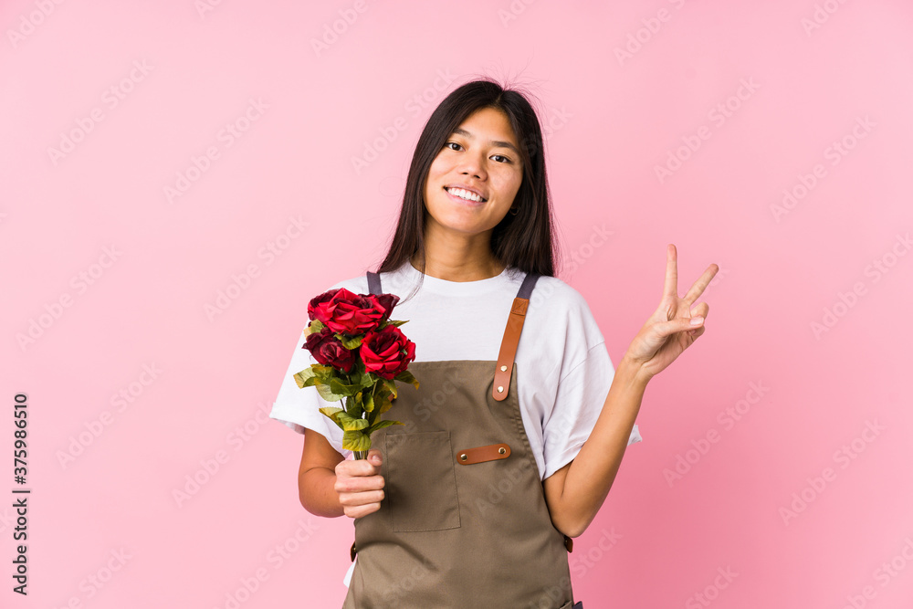 Young chinese gardener woman holding a roses isolated joyful and carefree showing a peace symbol with fingers.