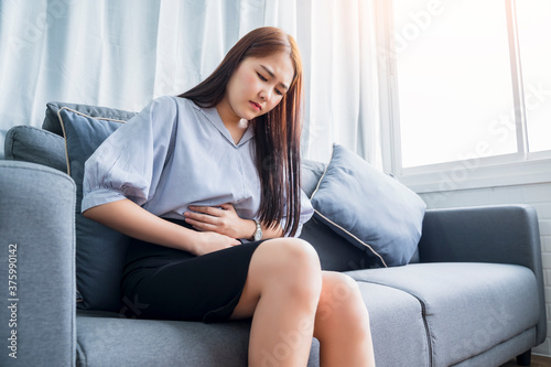 Asian mature woman having monthly period menstrual menstruation cycle pain symptoms problems, feeling painful ache in her belly stomach sitting on sofa couch modern living room formal business wear