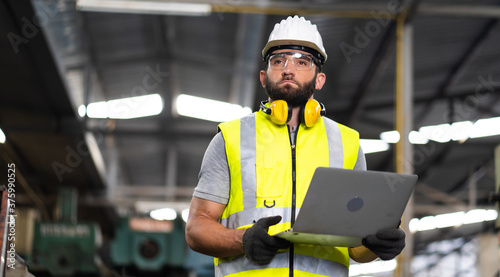 Young caucasian engineering man in Hard Hat Wearing Safety Jacket working on laptop computer in Heavy Industry Manufacturing Facility.